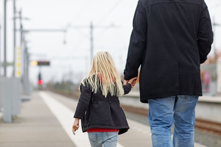 A young girl with blonde hair holds her parent's hand as they walk down a train platform